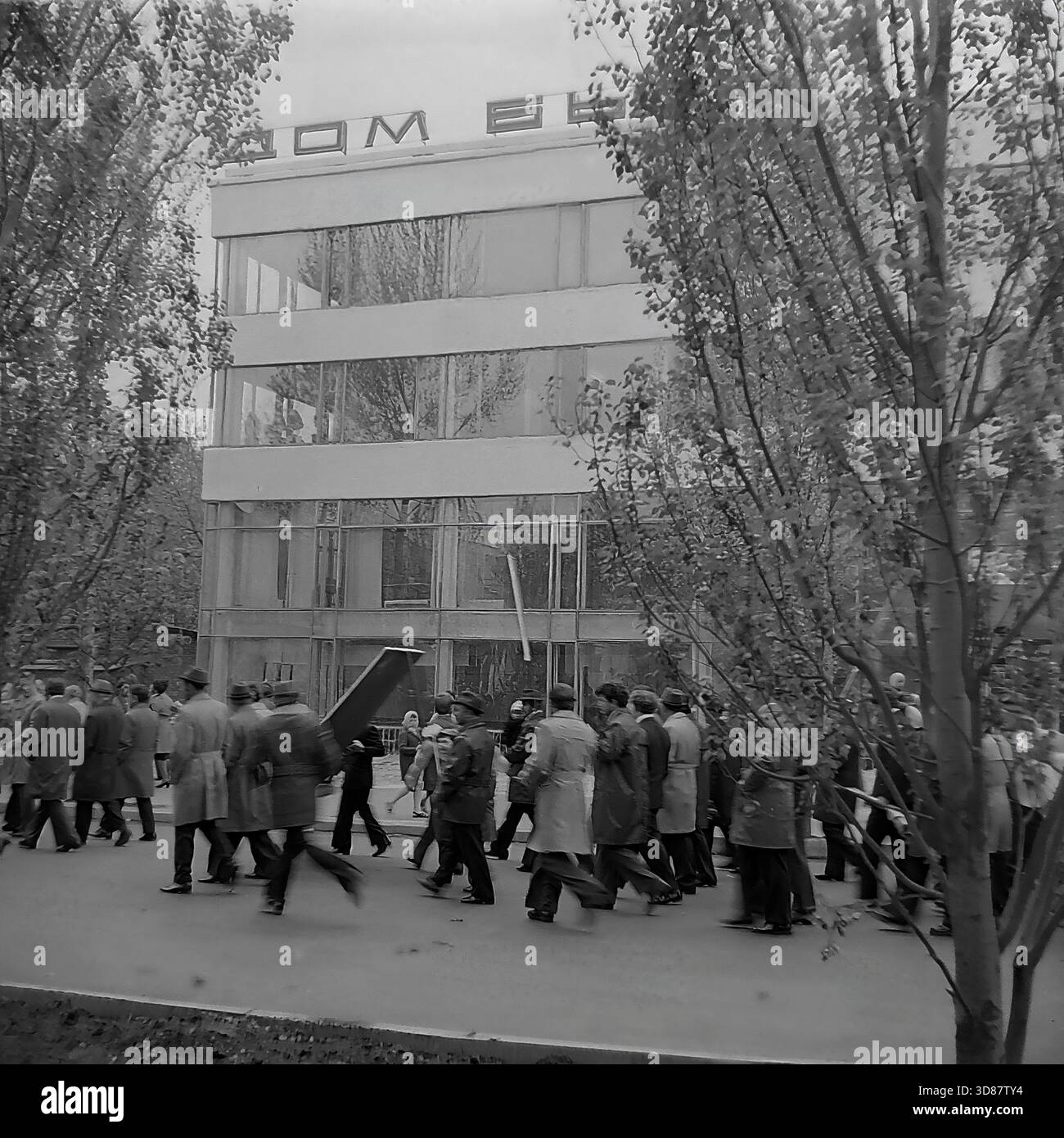 Slavyansk, Ucraina, URSS - 1970s: Una sfilata di manifestazioni festive in via Karl Marx (ora via Tsentralna). Le colonne dei residenti marciano oltre l'edificio modernista "Dom Byta" (House of Life Services). I partecipanti portano bandiere, palloncini, striscioni con slogan come "abbiamo bisogno di pace!" e ritratti di leader sovietici, tra cui Leonid Brezhnev. La gente è vestita con cappotti autunnali. Questa foto d'archivio in bianco e nero cattura i rituali ideologici, la moda e l'atmosfera sociale della tranquilla città industriale di Donbas. Foto Stock