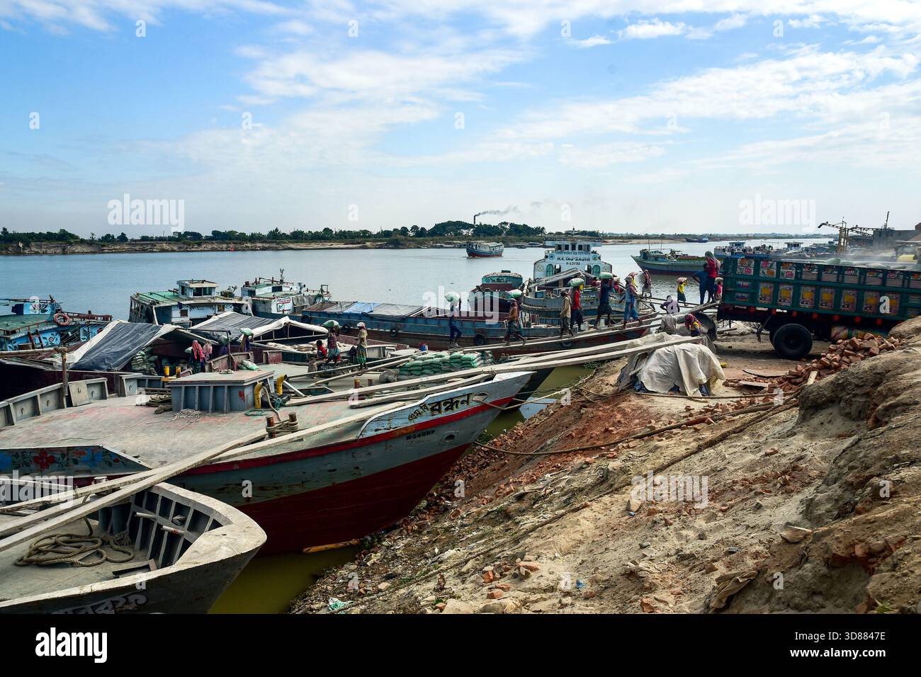 Porto sul fiume con barche colorate, camion e lavoratori lungo la costa durante le attività diurne Foto Stock