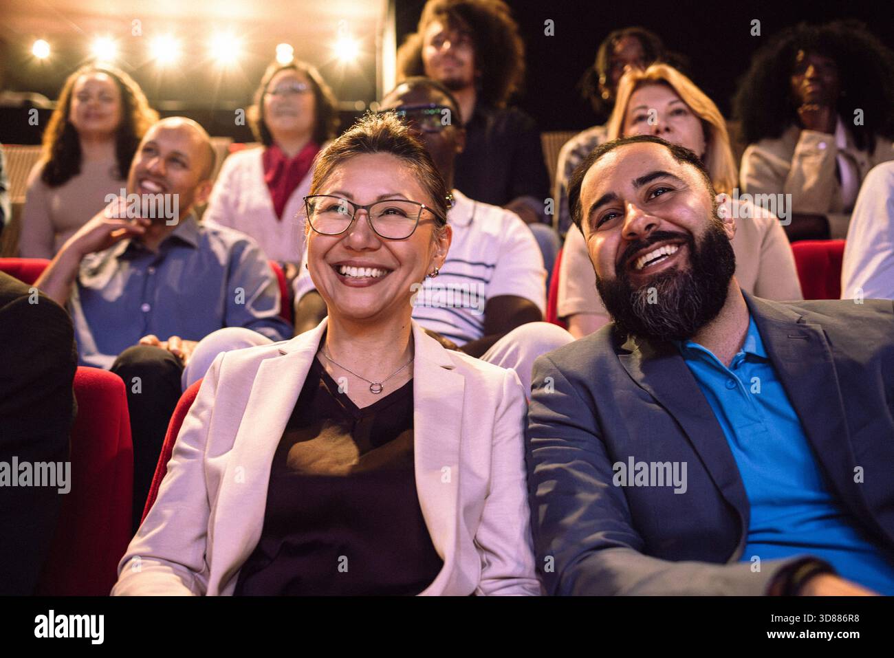 Amici sorridenti di sesso maschile e femminile che guardano il palco mentre sono seduti in un teatro illuminato Foto Stock