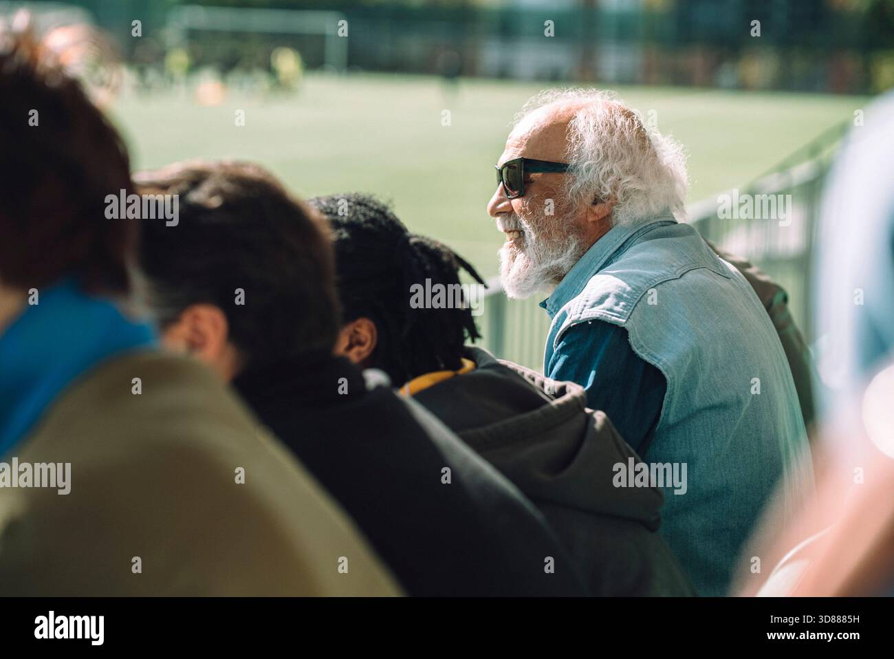 Vista laterale di un anziano sorridente che indossa occhiali da sole e guarda la partita mentre si siede davanti al pubblico allo stadio sportivo Foto Stock