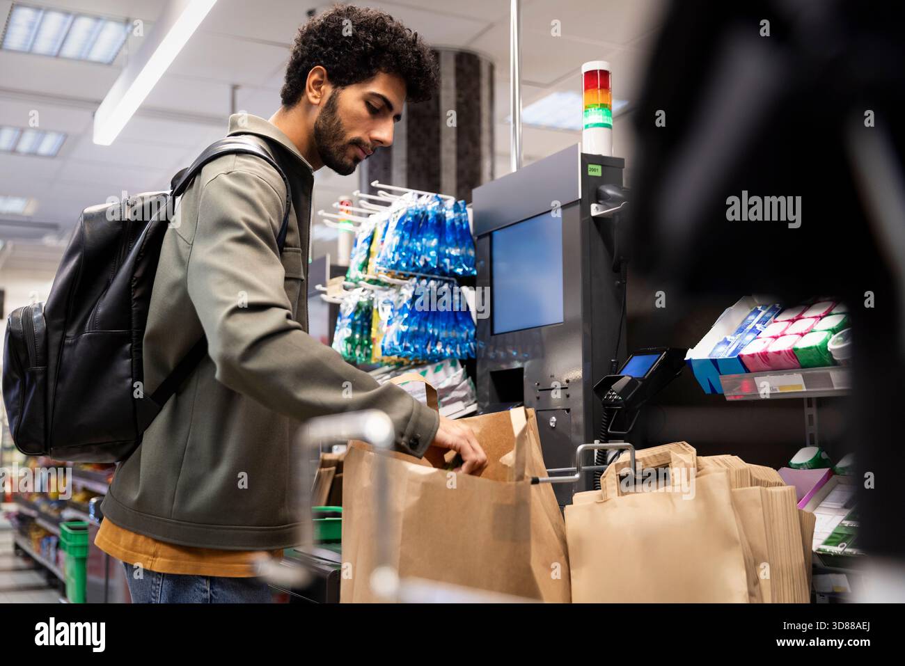 Uomo che trasporta zaino che rimuove la spesa dal sacchetto di carta al banco cassa self-service del supermercato Foto Stock