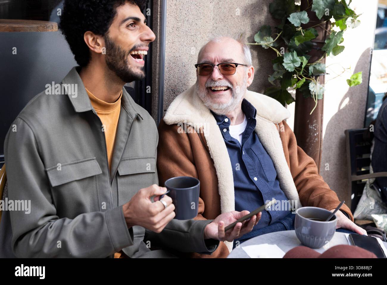 Uomo felice che tiene la tazza e lo smartphone mentre si diverte con il padre in un bar Foto Stock