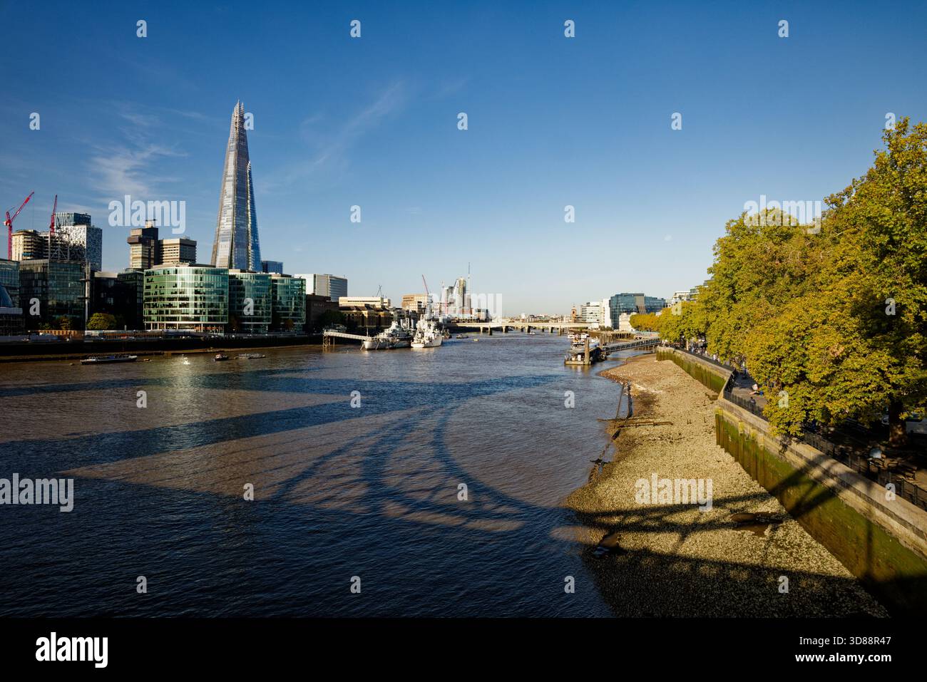 L'ombra del Tower Bridge sul fiume Tamigi con alberi lungo il fiume e l'imponente grattacielo Shard sulla South Bank, Londra, Inghilterra. Foto Stock