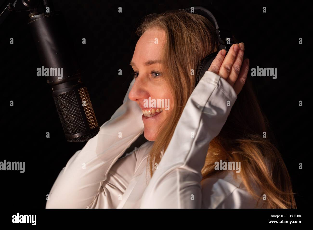 Donna dai capelli lunghi, che indossa una camicia bianca, si trova in uno studio di registrazione, regola le cuffie e sorride, immersa nel processo di creazione della musica Foto Stock