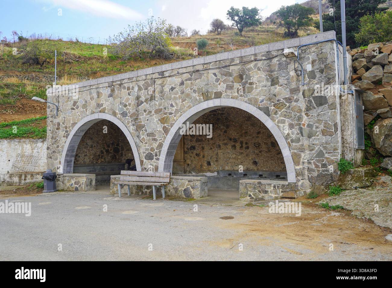Tradizionale fontana in pietra del villaggio di Melampes, Creta, caratterizzata da due grandi archi in pietra, grezzo muratura in pietra e una semplice panca in legno Foto Stock