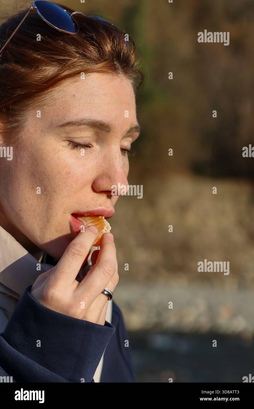 Donna che porta in bocca un succoso segmento di mandarino vicino al fiume calmo, sotto la calda luce del sole autunnale Foto Stock
