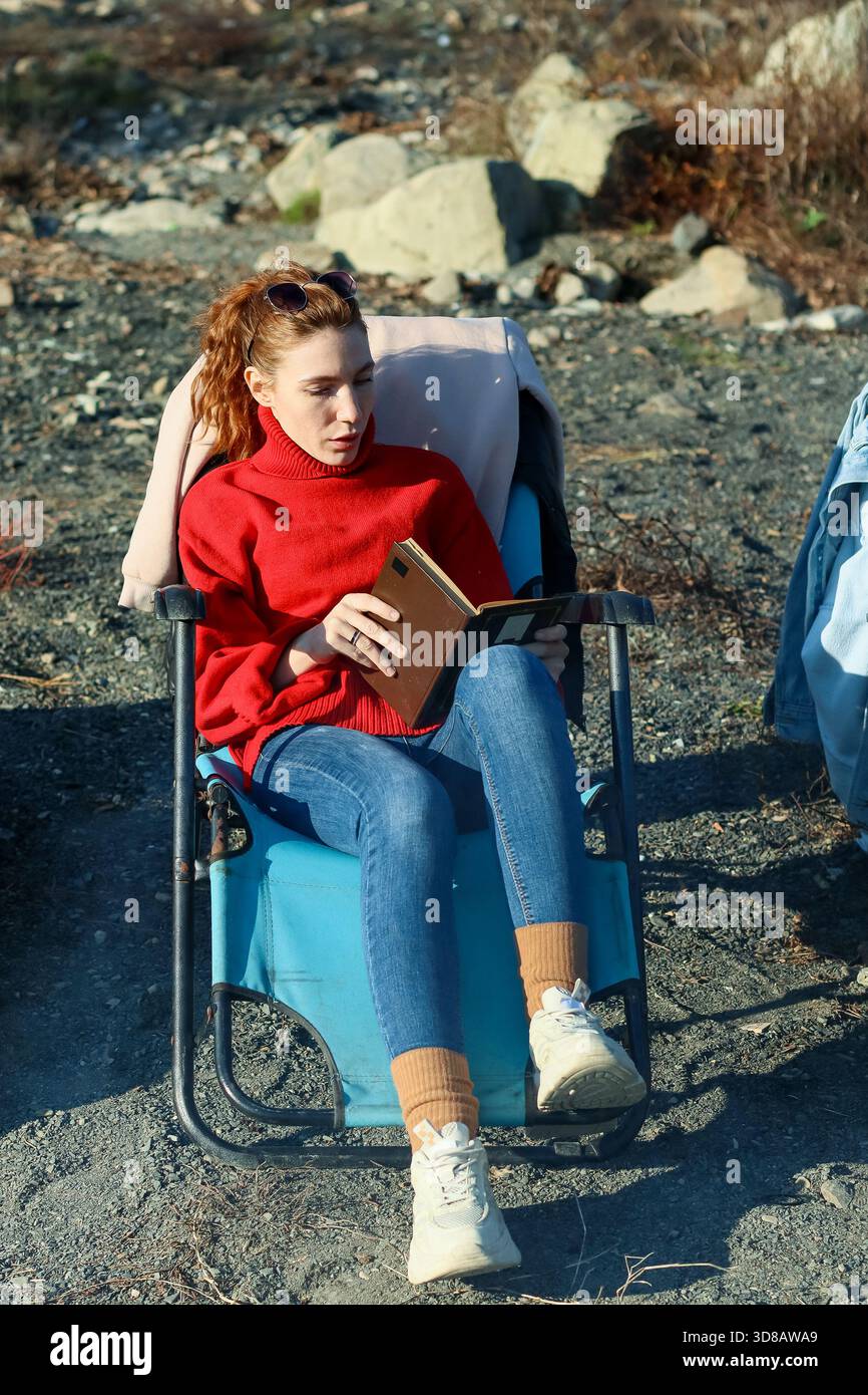 Giovane donna adulta con lentiggini e coda di cavallo, con maglione rosso e jeans, legge il libro mentre è seduta su una sedia blu su un terreno roccioso Foto Stock