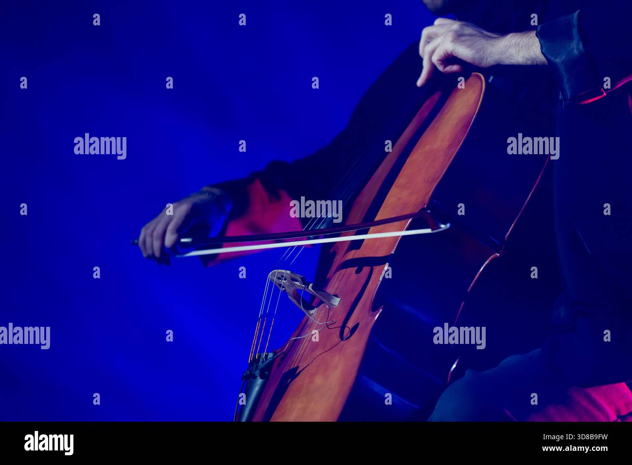 Musicista che si esibisce su un violoncello di legno sotto una vivace illuminazione blu del palcoscenico con chiara attenzione al movimento dell'arco e alla postura espressiva della mano durante un conce dal vivo Foto Stock