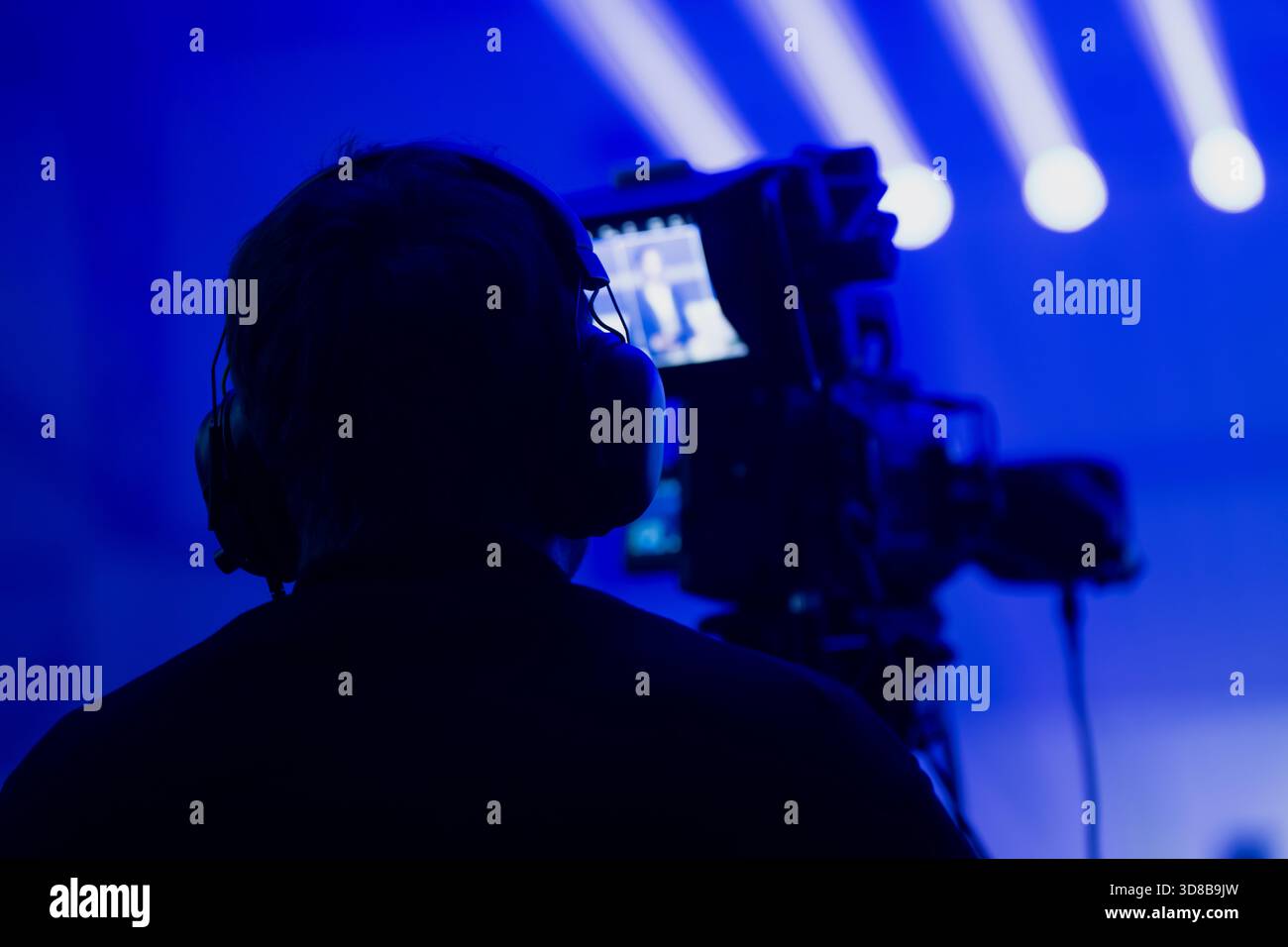 Scena di eventi dal vivo con un operatore che cattura riprese in condizioni di luce blu intenso, mentre potenti fasci di luce illuminano lo sfondo Foto Stock
