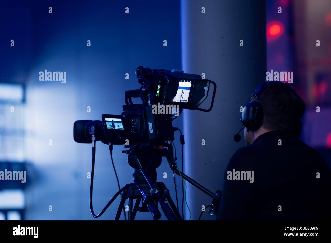Operatore video di eventi dal vivo che utilizza un sistema di telecamere broadcast con treppiede, cuffie di comunicazione e monitor sulla telecamera all'interno di un locale di produzione interno Foto Stock