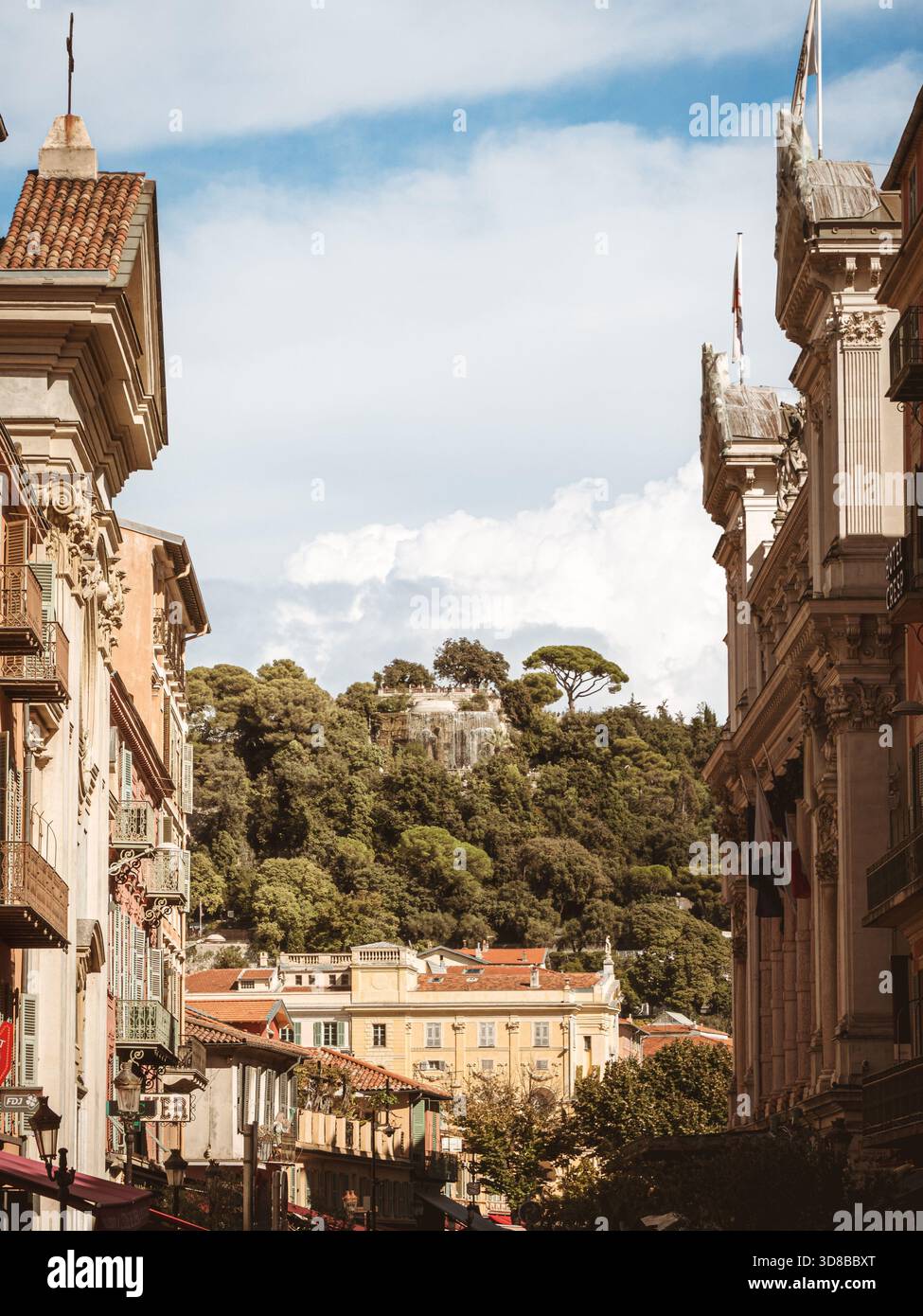 Una strada affascinante a Nizza, Francia, conduce lo sguardo verso la lussureggiante vegetazione e l'iconica cascata della Collina del Castello, incorniciata da eleganti edifici storici Foto Stock