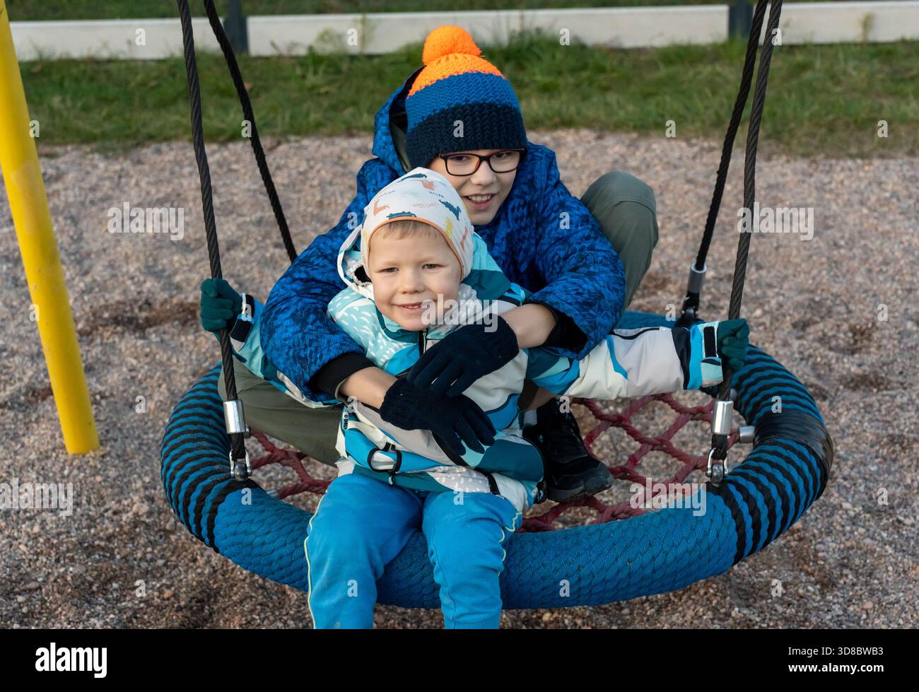 Due fratelli in giacche invernali seduti insieme su un nido rotondo al parco giochi, un ragazzo più grande abbraccia il fratello più piccolo nella fredda giornata autunnale. Concetto di Foto Stock