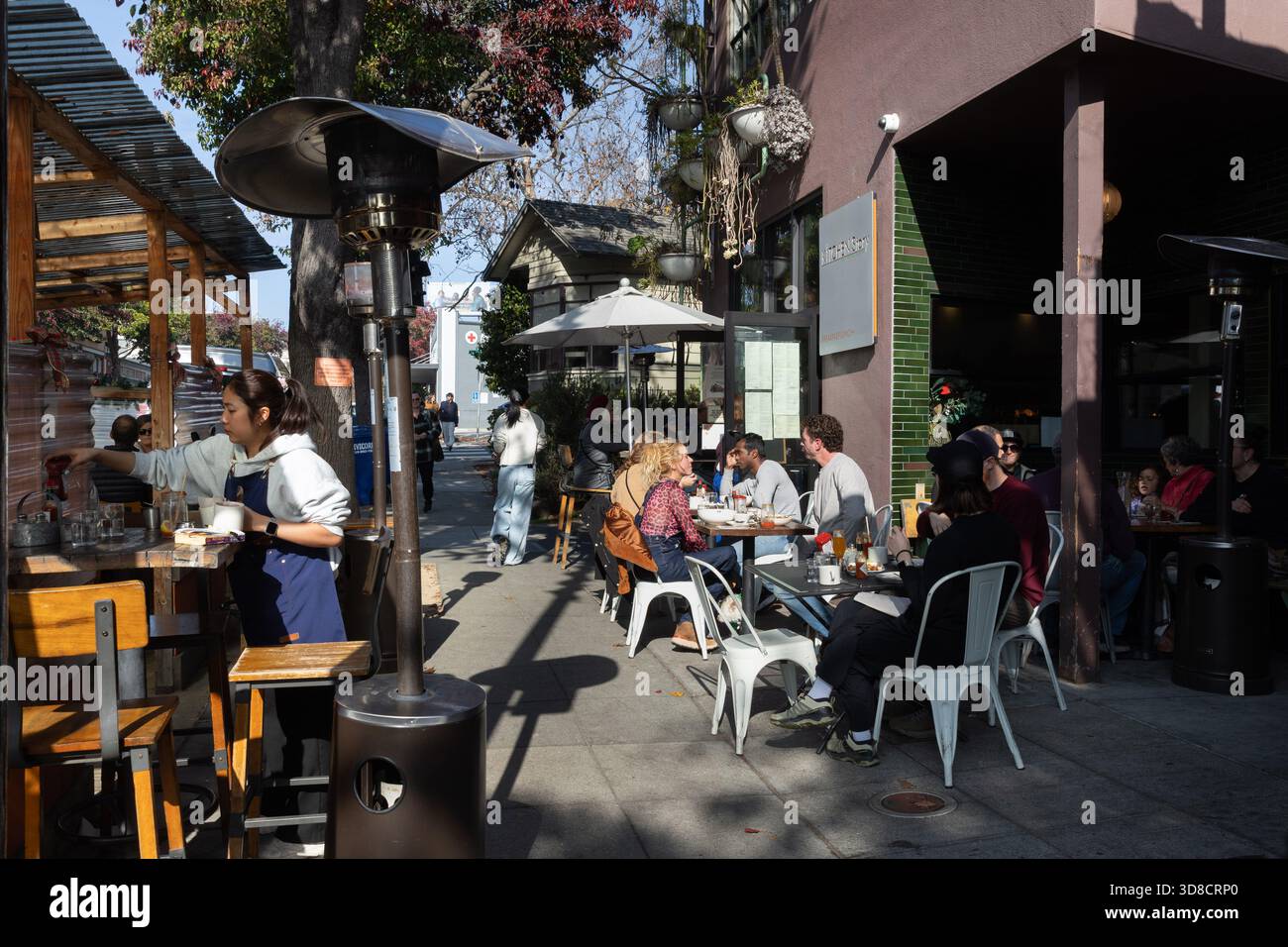 I clienti potranno gustare un pasto pomeridiano soleggiato ai tavoli accanto a un affollato ristorante Oakland, mentre il personale lavora nelle vicinanze lungo la strada alberata. Foto Stock