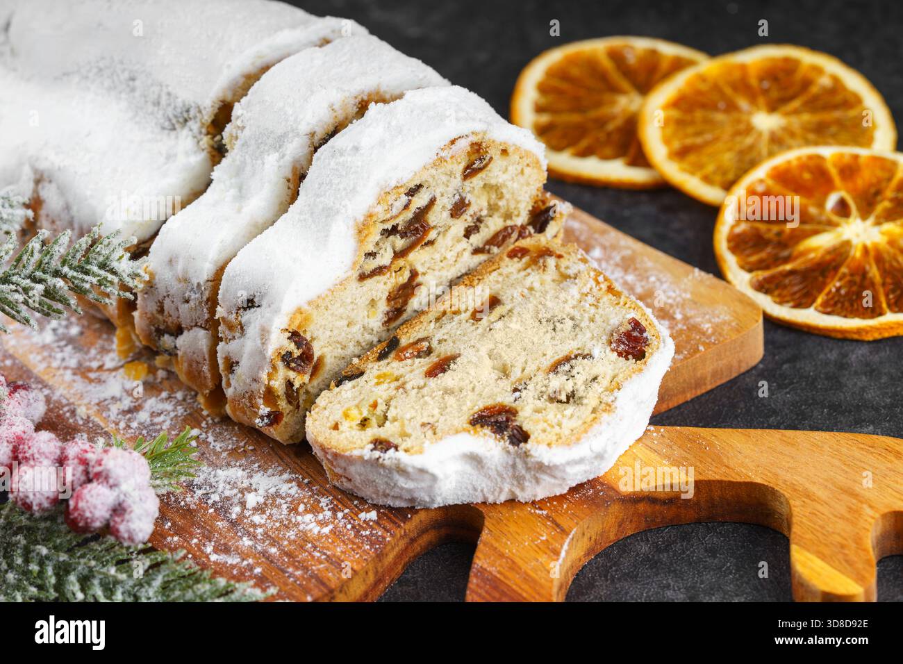 Tradizionale panino natalizio, pane di frutta dolce tagliato su tavola di legno, spolverato con zucchero a velo su fondo nero testurizzato. Spezie, frutta secca Foto Stock