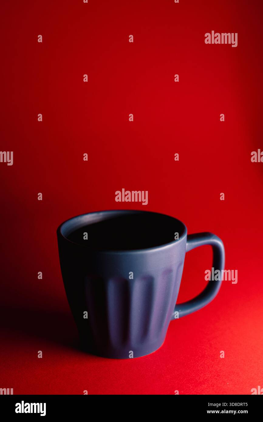 Una tazza blu opaca si trova su una superficie rossa vivace, creando un forte contrasto minimalista in un ambiente da studio pulito. Foto Stock