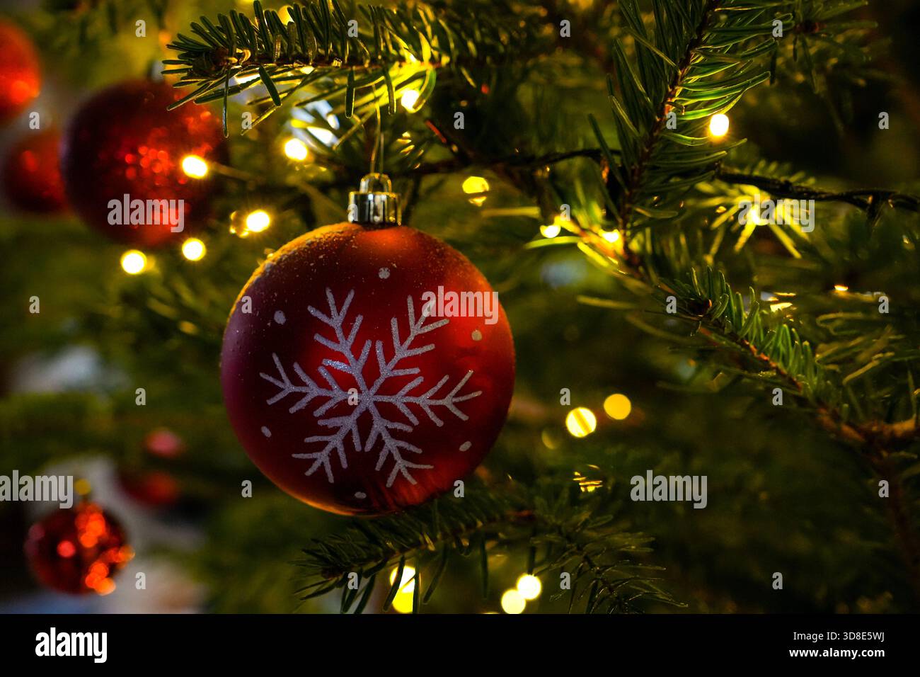 Primo piano di un ornamento natalizio rosso con un fiocco di neve bianco appeso su un albero di Natale illuminato. Le calde luci delle fate creano un magico effetto bokeh e. Foto Stock