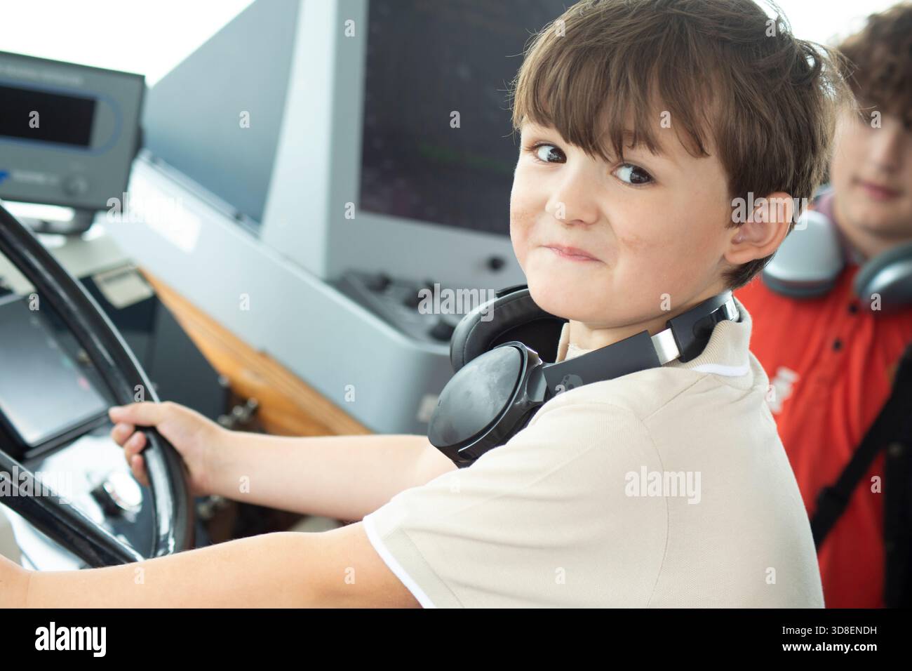 Bambino che tiene in mano una ruota di controllo all'interno di un'imbarcazione o di un'ambientazione storica, sorridendo verso la macchina fotografica durante una visita didattica e un'esperienza pratica di apprendimento Foto Stock