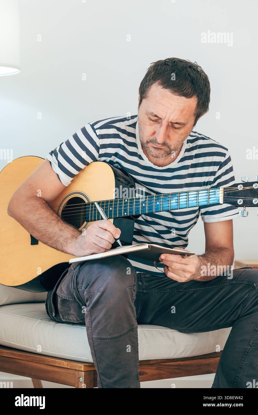 Chitarrista maturo che scrive una canzone a mano mentre si pratica la chitarra acustica al chiuso, concentrandosi sulla creazione di musica. Messa a fuoco selettiva. Foto Stock