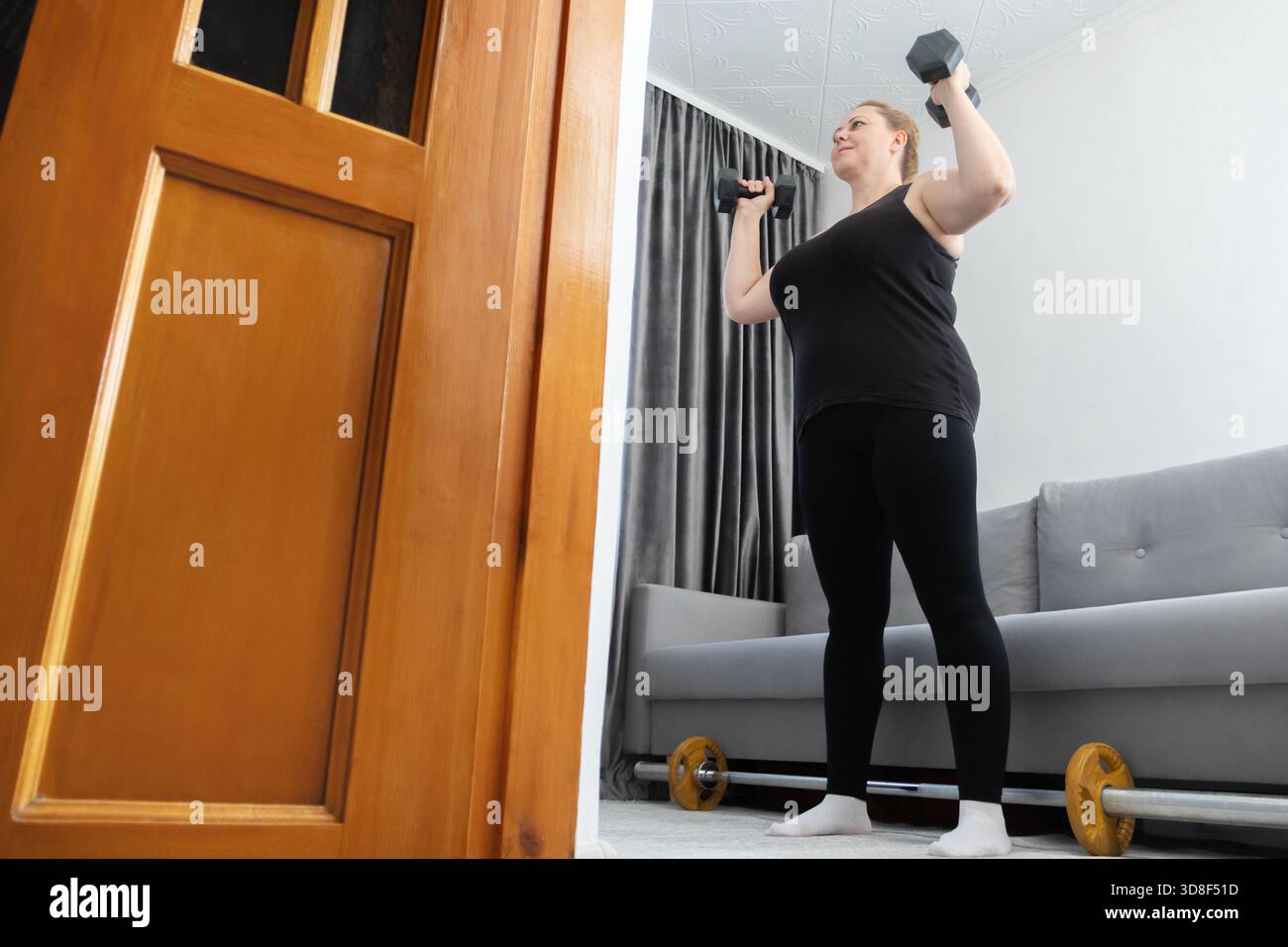 Inoltre, una donna di taglia taglia taglia si allena seriamente con i manubri nel suo salotto di casa. per promuovere la salute, il fitness e la positività del corpo. Foto Stock