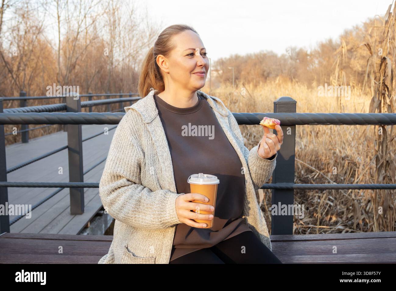 Donna di mezza età felice seduta all'aperto per una pausa con ciambelle e caffè. per temi di svago, stile di vita, positività del corpo e relax. Foto Stock