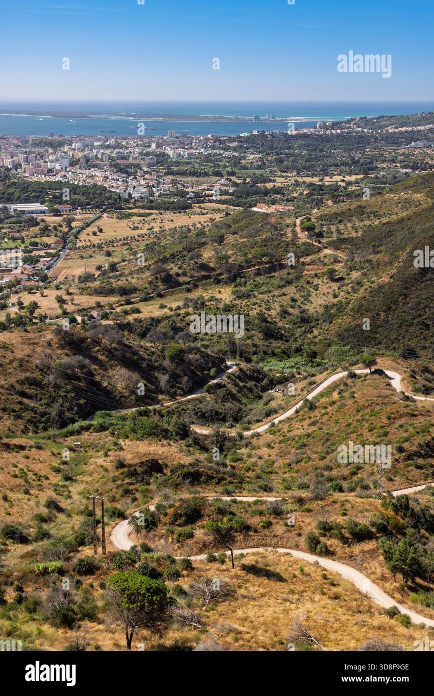 Vista verticale ad angolo alto dal Parco naturale di Arrábida, Portogallo. Una strada tortuosa serpeggia attraverso colline asciutte, con la città di Setúbal e la costa. Foto Stock
