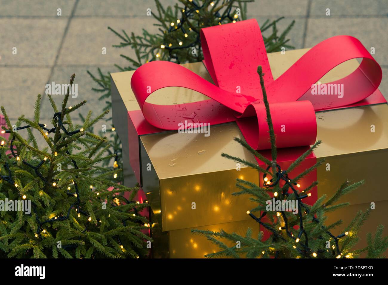 Una grande scatola regalo dorata con un nastro rosso si trova tra piccoli alberi di Natale decorati con luci fiabesche Foto Stock