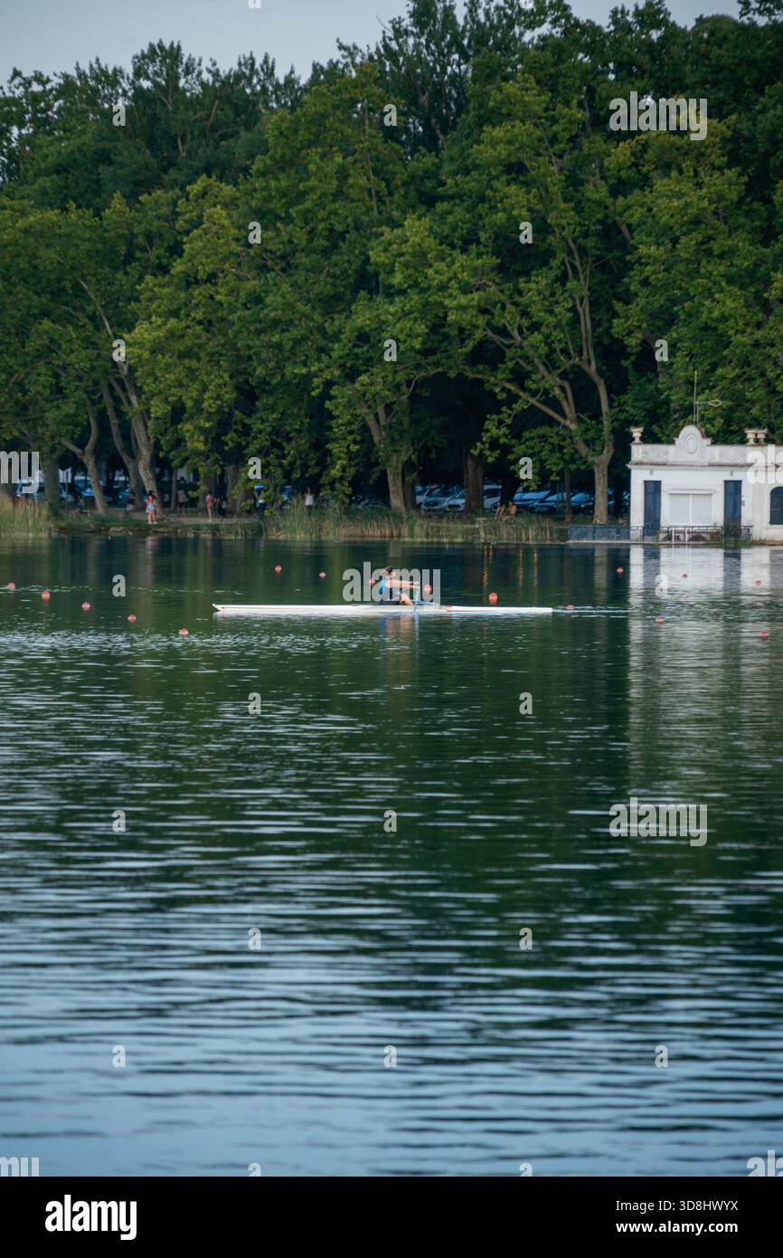 Un canottaggio si allena su una barca sulle calme acque del lago Banyoles, con la storica Pesqueres e la foresta costiera sullo sfondo Foto Stock