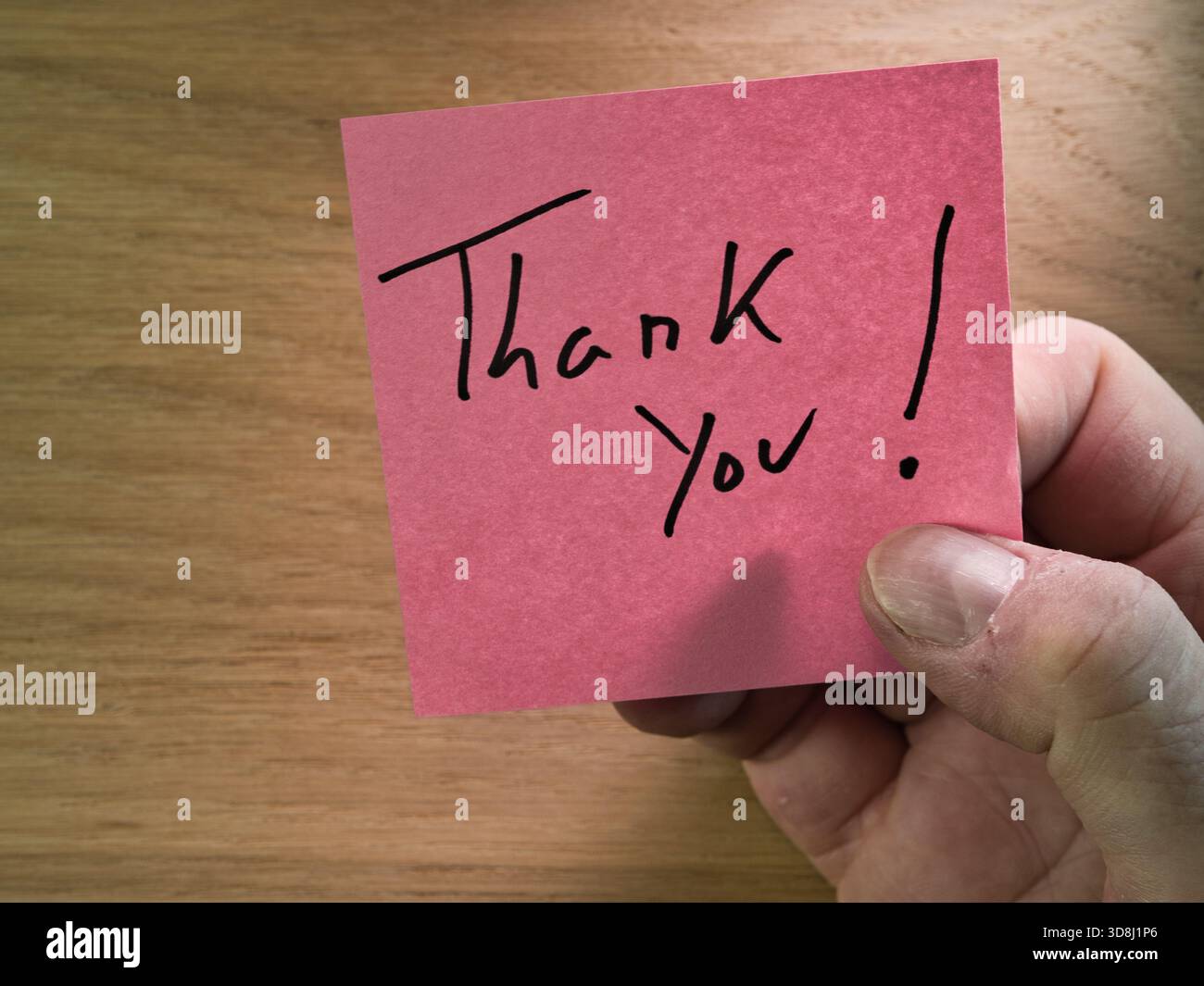 Nota di ringraziamento tenuta a mano su legno - mano che tiene una nota adesiva rosa vivace con "Grazie!" scritto a mano, su un caldo sfondo di legno. A c Foto Stock