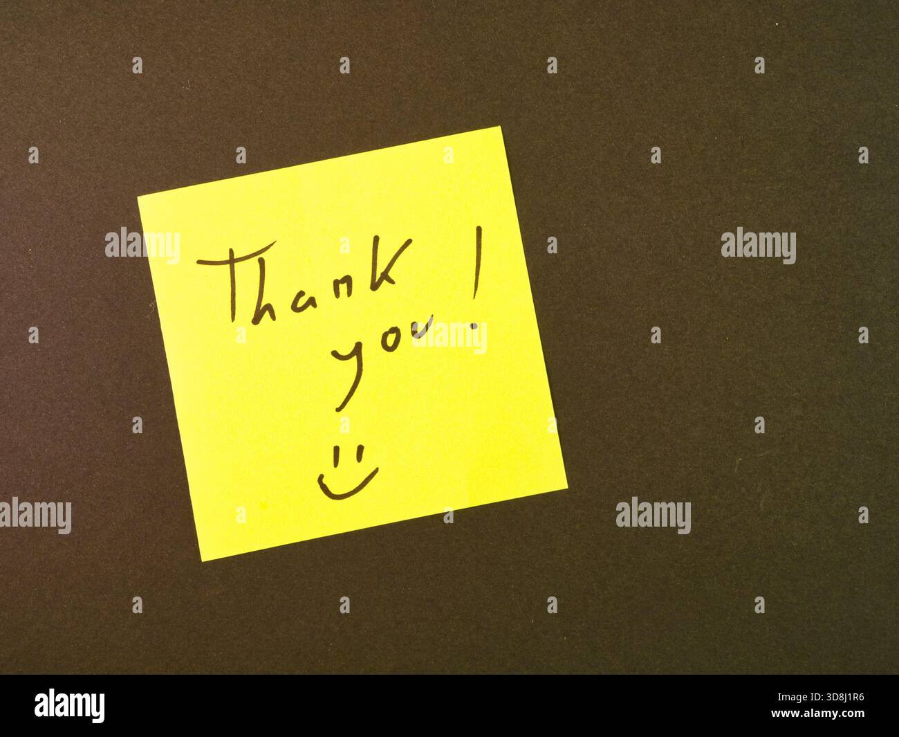 Nota di ringraziamento con la faccia Smiley - Una nota adesiva gialla brillante mostra un sentito "Grazie!" messaggio e una faccia sorridente contro un bac scuro e strutturato Foto Stock