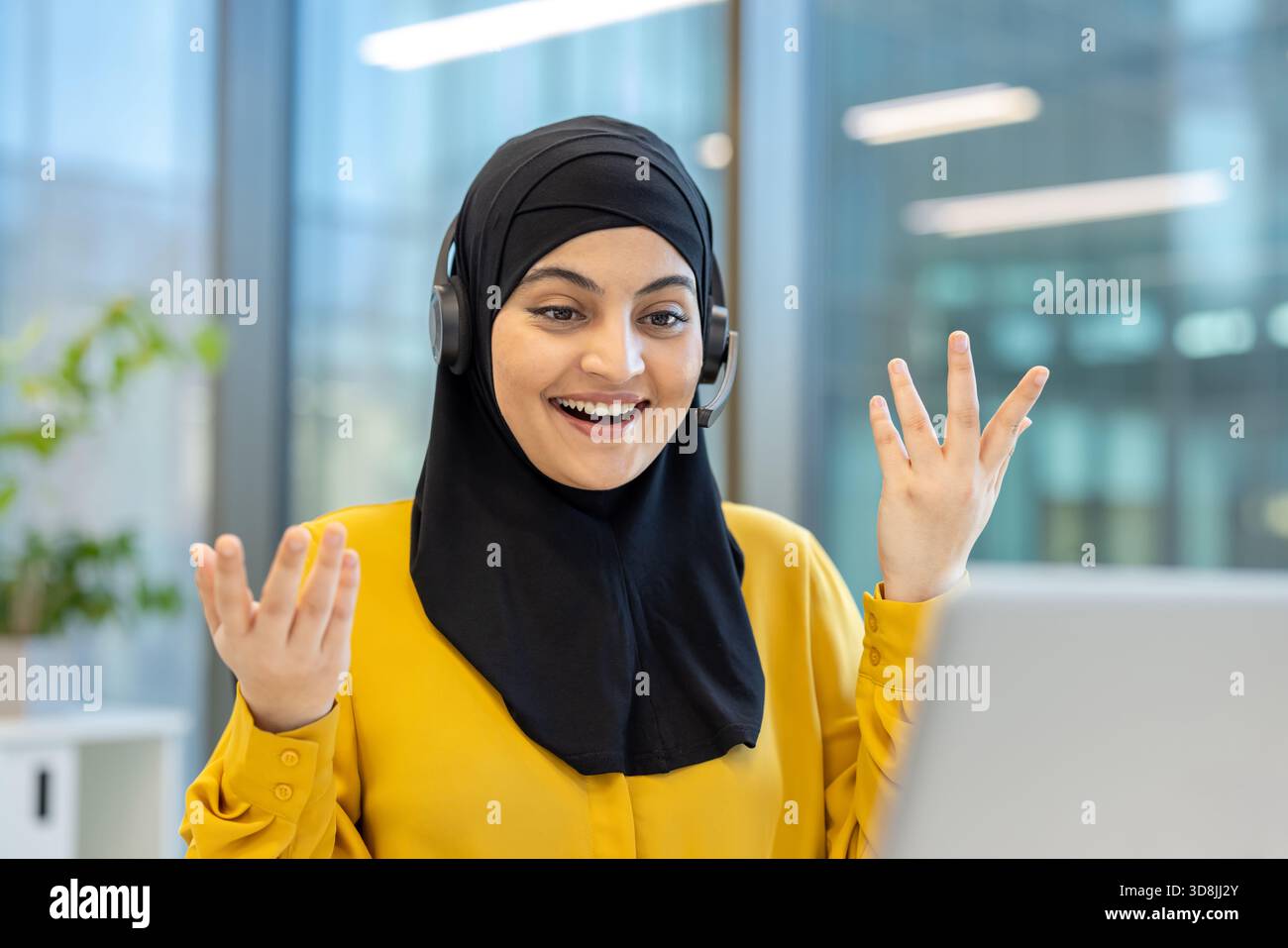 Donna musulmana che indossa un hijab nero e cuffie, sorridente e gestante mentre partecipa a una videochiamata online dal suo ufficio moderno, comunica e collabora virtualmente Foto Stock
