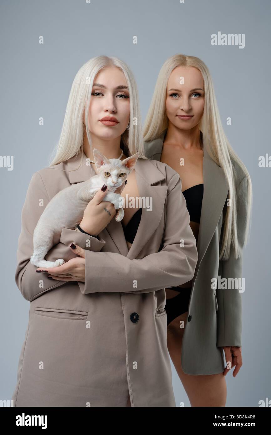 Due donne con i capelli lunghi stanno insieme, una con un cane piccolo, entrambe con blazer alla moda in un ambiente interno moderno. || modello rilasciato Foto Stock