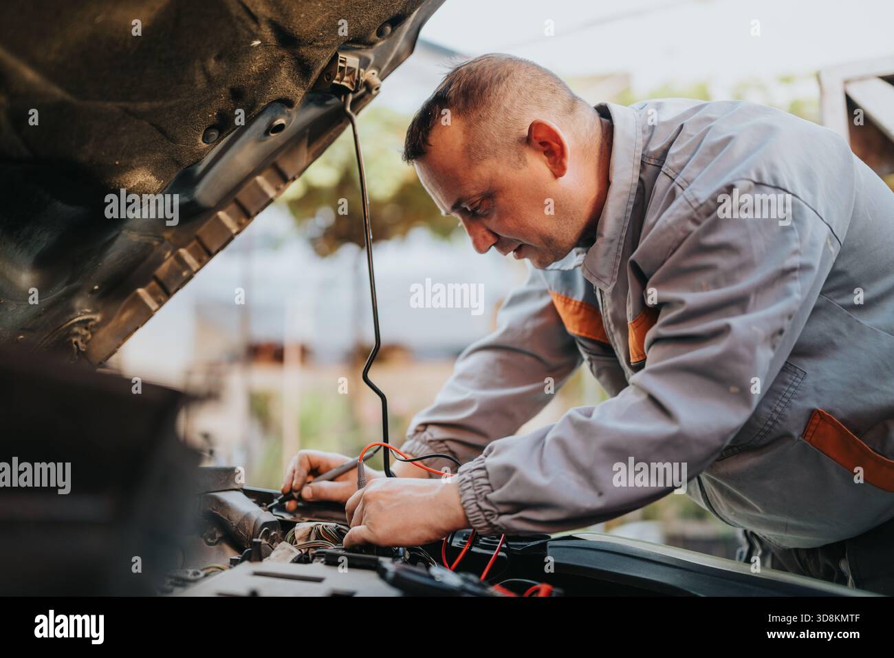 Meccanico maschio che ripara il motore dell'auto sotto il cofano all'aperto, concentrandosi sul cablaggio elettrico e sulla diagnostica Foto Stock