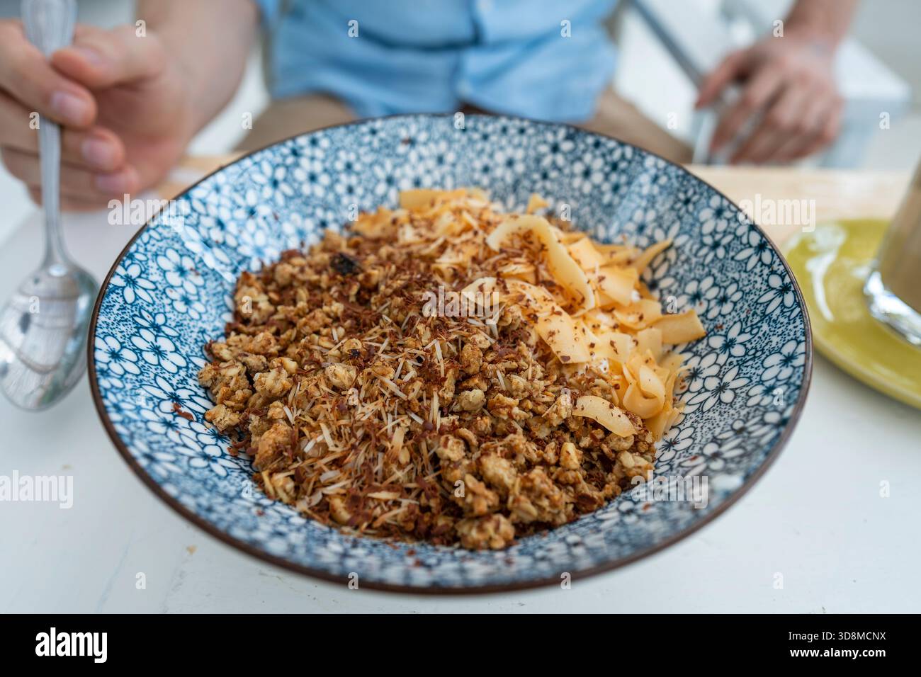Ciotola con granuli e patatine di cocco e cioccolato per una colazione sana. Mano maschio con un cucchiaio e un piatto con muesli in un caffè al mattino, c Foto Stock