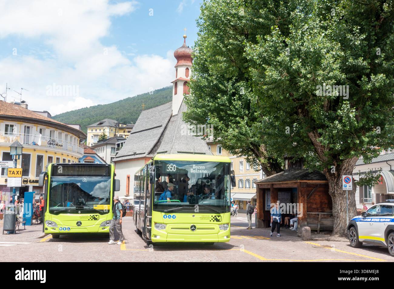 Vivace scena estiva in Piazza SantAntonio, Dolomiti. Autobus verdi in una fermata trafficata con persone che si imbarcano, una torre della chiesa e edifici alpini sullo sfondo Foto Stock