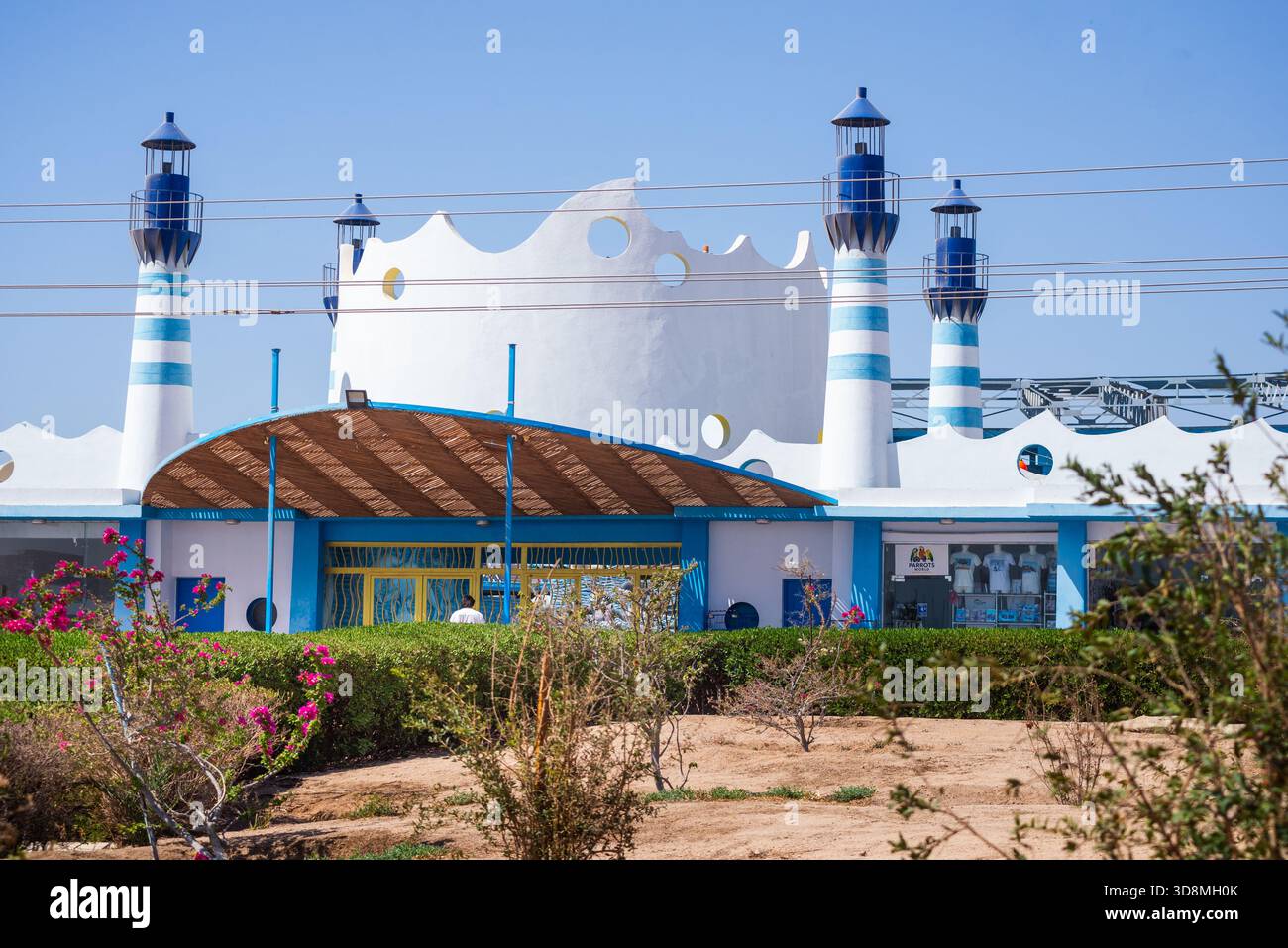 Il Delphinarium di Hurghada sorge sotto un cielo azzurro, torri bianche e blu che si innalzano su siepi e piante desertiche, con linee elettriche che tagliano acr Foto Stock