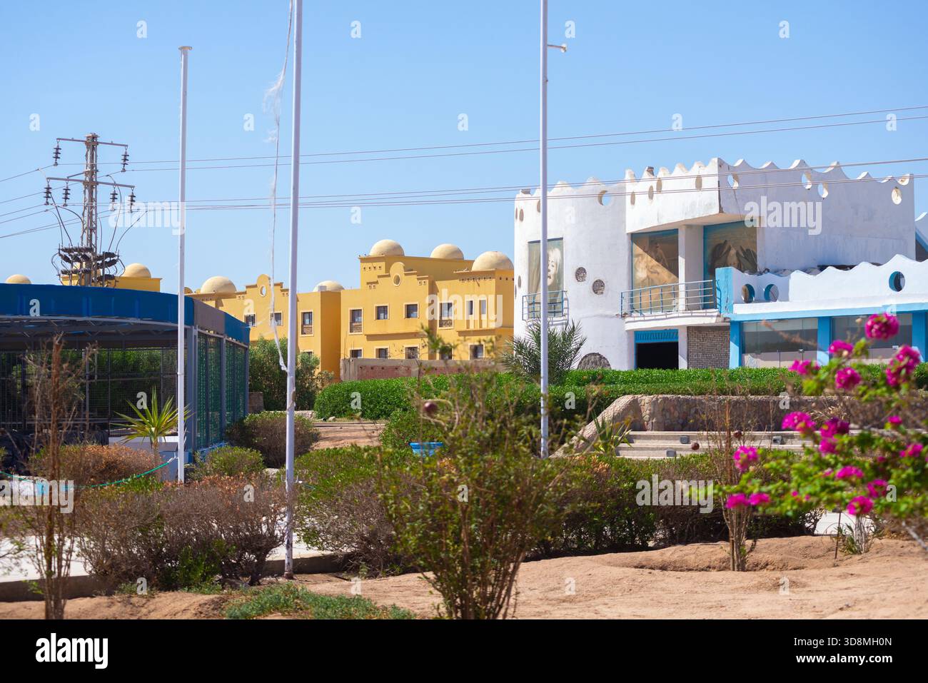Edifici in Delphinarium bianchi e blu si trovano accanto a strutture a cupola gialla a Hurghada, incorniciate da siepi, terreno desertico, arbusti fioriti, flagpole e. Foto Stock