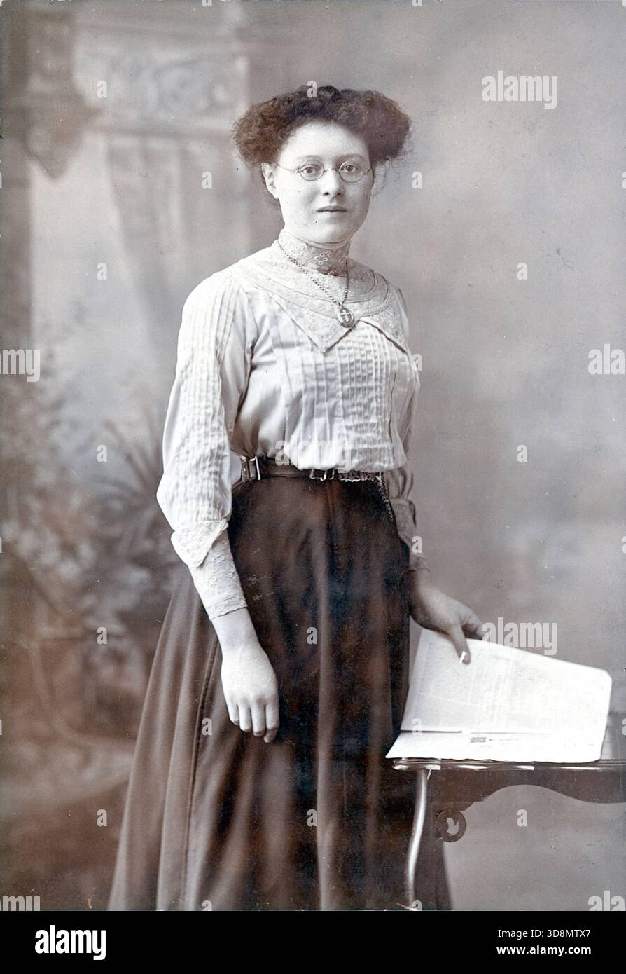 Un ritratto in studio di Henry Bown di New Kent Road e altre località di Londra. Una persona sta accanto a un tavolo ornato con un libro aperto, incorniciato da uno sfondo dipinto di architettura classica e fogliame. La loro blusa a pieghe, la gonna scura, i capelli voluminosi e gli occhiali evocano la moda e il portamento erudito dei primi anni del XX secolo all'interno di una tradizione fotografica formale. Da un album fotografico del tardo vittoriano a quello del primo edoardiano, che presentava molte carte di albume e carte de visites, fornendo una finestra sugli stili e le mode dell'epoca. Foto Stock