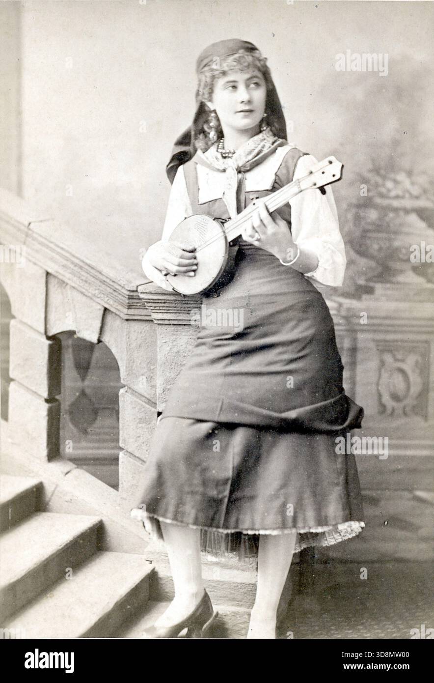 Florence St. John si erge su una scala di studio, tenendo un banjo in costume per il ruolo del protagonista in Madame Favart allo Strand Theatre, 1879. La sua gonna lunga, la blusa e il velo riflettono lo stile teatrale dell'epoca. Lo sfondo dipinto e la scena musicale evocano la scena vittoriana, la rappresentazione del personaggio e il fascino fotografico della London Stereoscopic Company. Da un album fotografico del tardo vittoriano a quello del primo edoardiano, che presentava molte carte di albume e carte de visites, fornendo una finestra sugli stili e le mode dell'epoca. Foto Stock