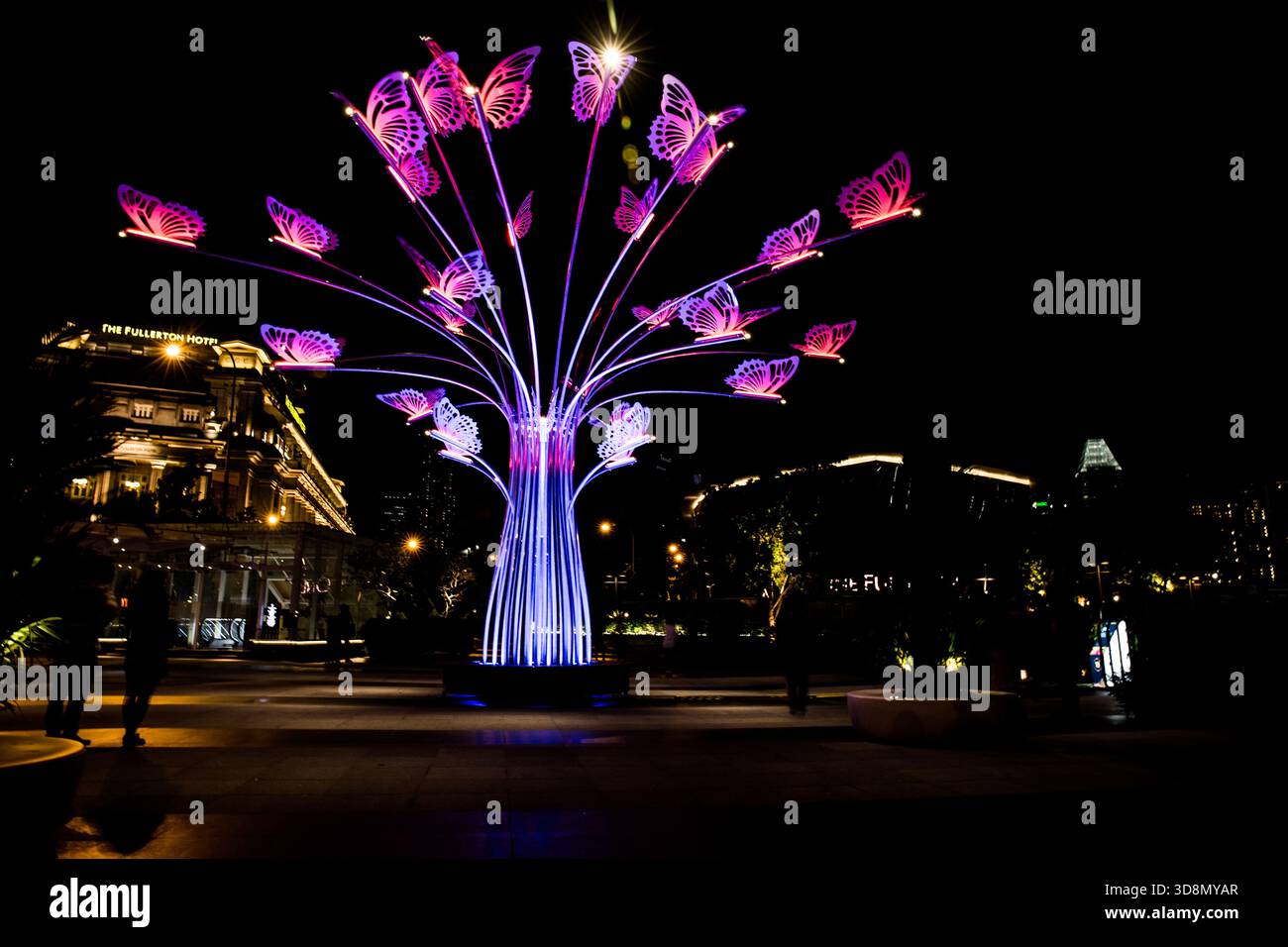 La scultura illuminata dell'albero delle farfalle si illumina sotto il cielo notturno, proiettando una luce colorata sulla piazza urbana. Foto Stock