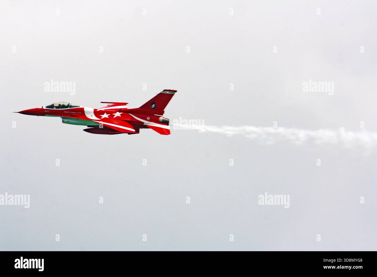 Un caccia rosso e bianco F-16 vola attraverso un cielo nuvoloso, lasciando una pista di fumo bianca, che simboleggia la precisione e la velocità militari Foto Stock