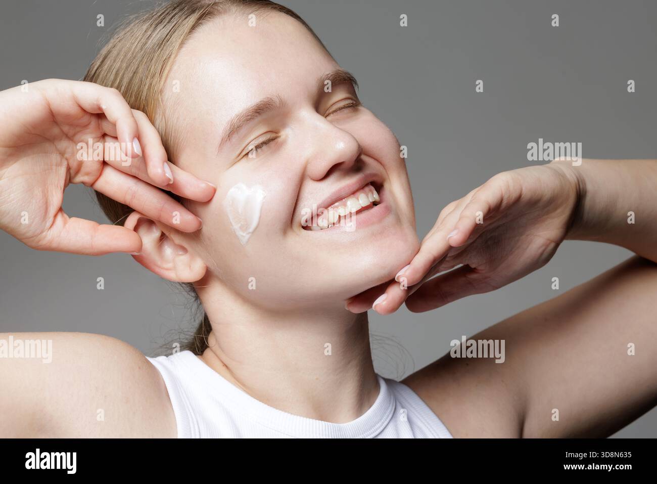 Donna sorridente con gli occhi chiusi e cuore di crema idratante sulla guancia che mostra gioia e delicata cura quotidiana del viso Foto Stock