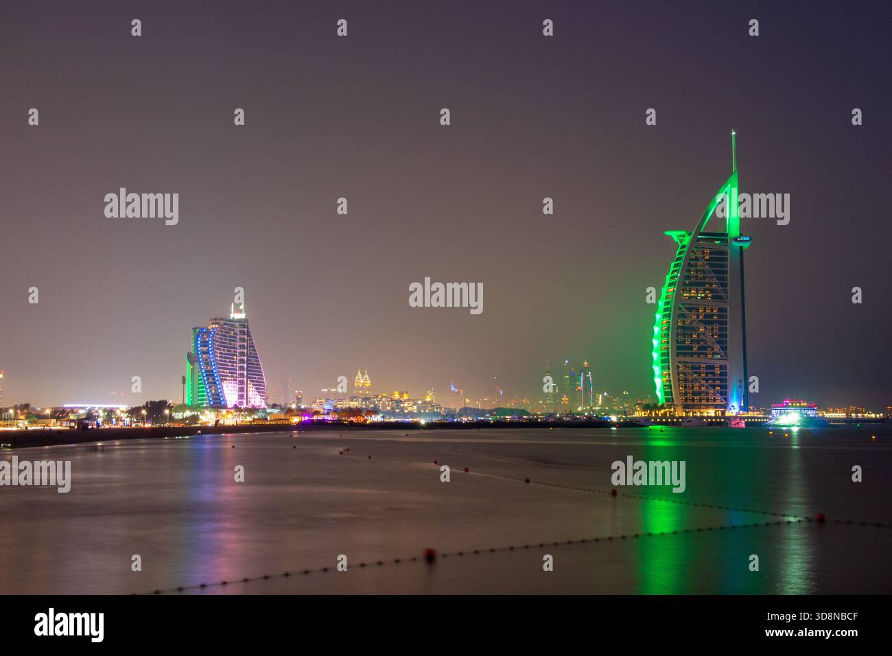 Vista del Burj al Arab illuminato e del Jumeirah Beach Hotel che si riflette sulle tranquille acque, proiettando un vibrante bagliore contro il cielo scuro, Dubai, Foto Stock
