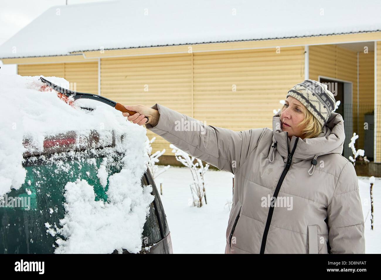 La spazzolatura rimuove la neve profonda dal tetto dell'auto mentre la donna indossa una calda giacca invernale e un cappello in maglia sgombra il veicolo vicino a una parete di legno in un giardino innevato. Foto Stock