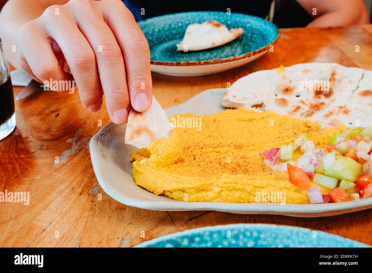 un uomo solleva il pane naan ricoperto di hummus al curry da un piatto condiviso su un tavolo del ristorante, mostrando lo spargimento giallo accanto alle verdure fresche tritate a. Foto Stock