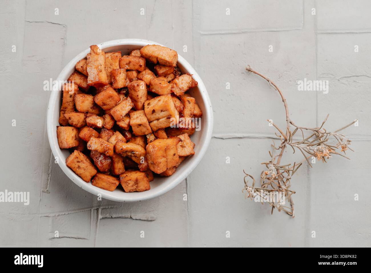 una ciotola piena di cubetti di tofu marinati, seduta su una superficie ruvida e leggera, che mostra il loro colore dorato e i bordi stagionati accanto a un piccolo ramoscello essiccato Foto Stock