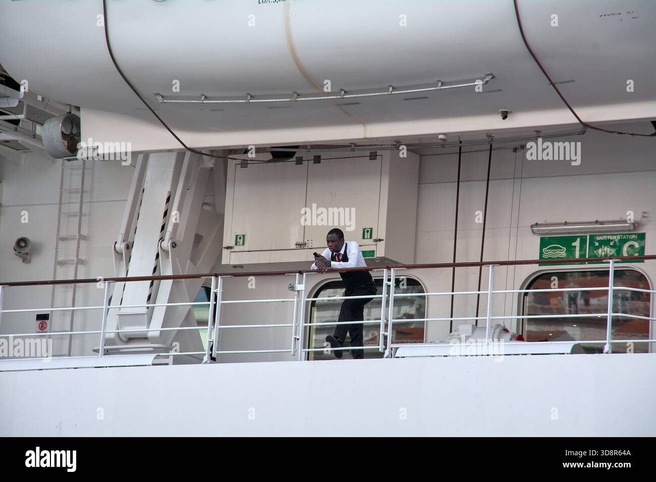 Dubrovnik. Croazia - 2 dicembre 2025: Cameriere o maggiordomo in uniforme (giubbotto e cravatta) si appoggia su una ringhiera sotto una scialuppa di salvataggio su una nave da crociera. Foto Stock