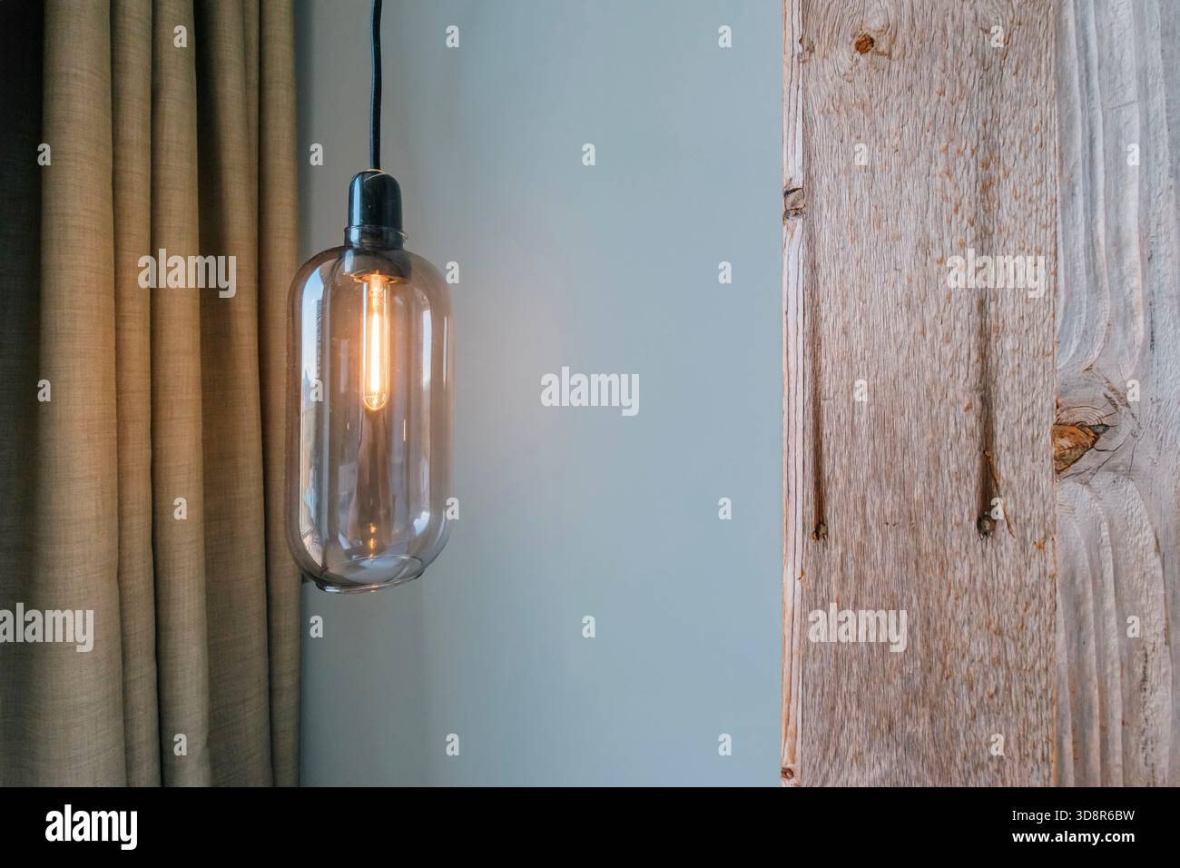 Lampada a sospensione con una lampadina a incandescenza accanto a una tenda beige testurizzata e un pannello in legno non finito Foto Stock