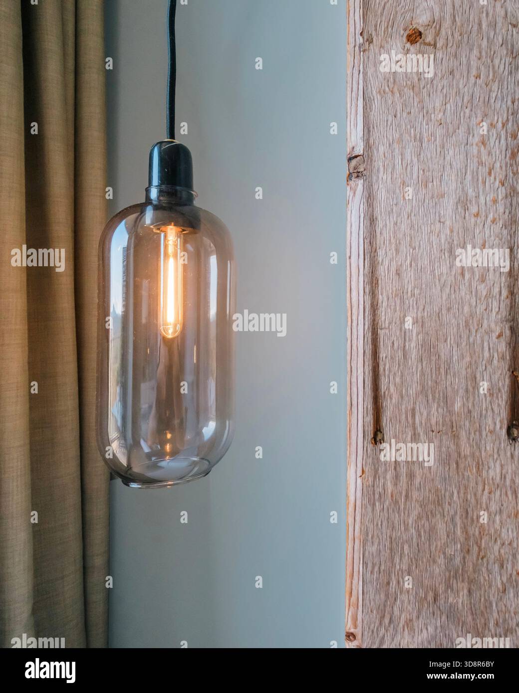 Primo piano di una lampada a sospensione in vetro con una lampadina a incandescenza, tra una tenda beige e un pannello in legno Foto Stock