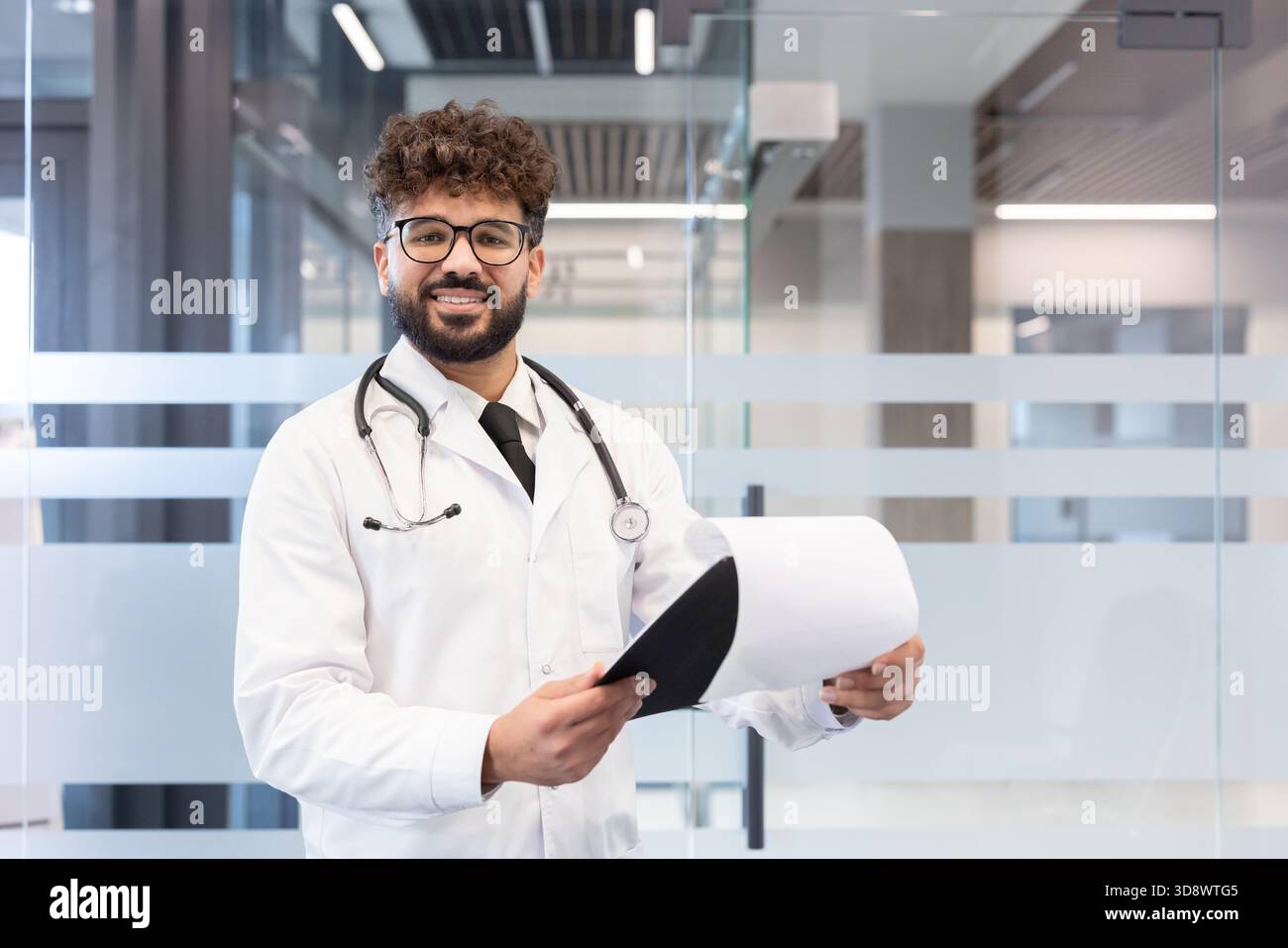 Giovane medico maschio che indossa un camice da laboratorio e uno stetoscopio, tenendo i documenti medici mentre si trova in una clinica moderna o in un ambiente ospedaliero, guardando la fotocamera e sorridendo Foto Stock
