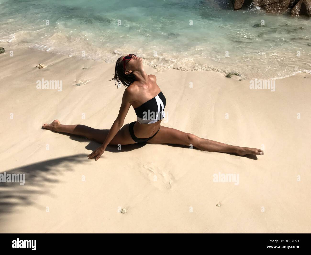Bella donna sportiva che si divide davanti, con una gamba in avanti e l'altra indietro, in bikini sulla spiaggia tropicale delle Seychelles. Modello femminile Foto Stock